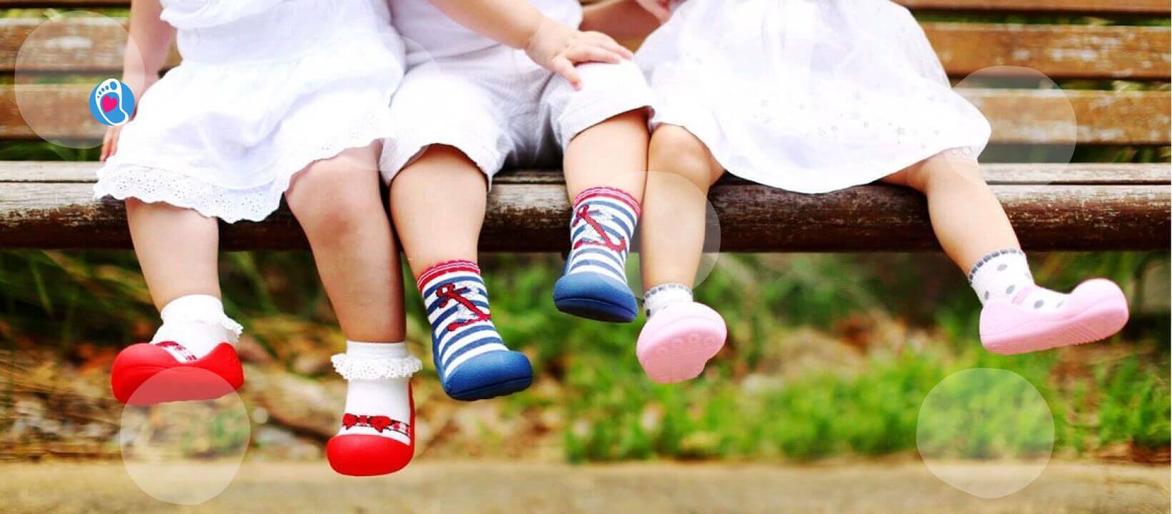Attipas–kolorowe-buciki-barefoot-dla-dzieci-na-letni-spacer Troje dzieci w kolorowych bucikach barefoot Attipas siedzi na drewnianej ławce w parku